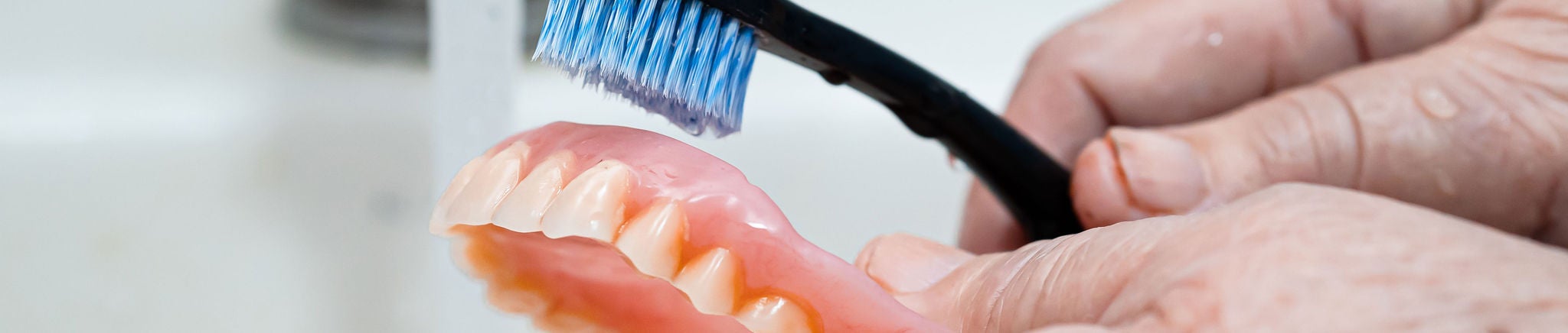 Someone holds their denture near faucent, with a toothbrush ready to clean it. 