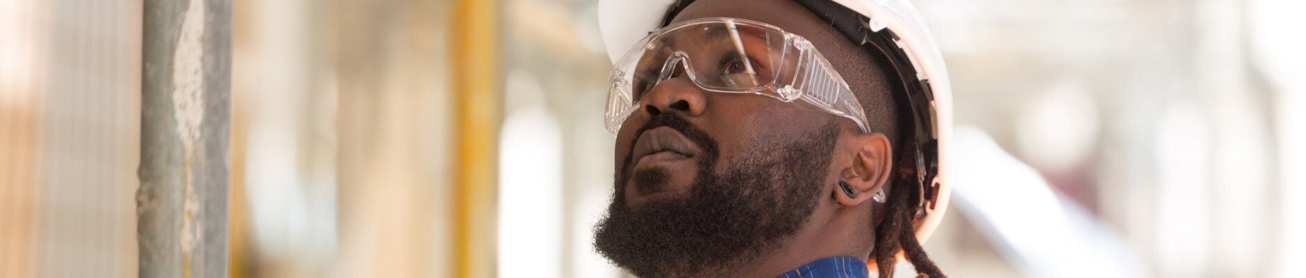 African American man wearing googles and a hard hat looking up at a construction site.