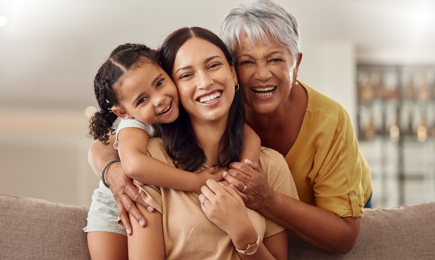 Young Hispanic woman sitting with older Hispanic woman and Hispanic child hugging her from behind.