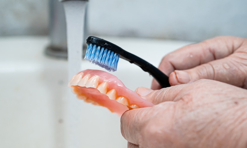 hands holding a black toothbrush with blue and white bristles cleaning dentures under a sink faucet